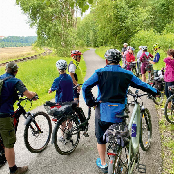 Eine Gruppe von Radfahrern mit Helmen macht eine Pause auf einem asphaltierten Radweg entlang einer Bahnlinie und Feldern, bereit für den nächsten Teil ihrer Radreise mit dem Bus.