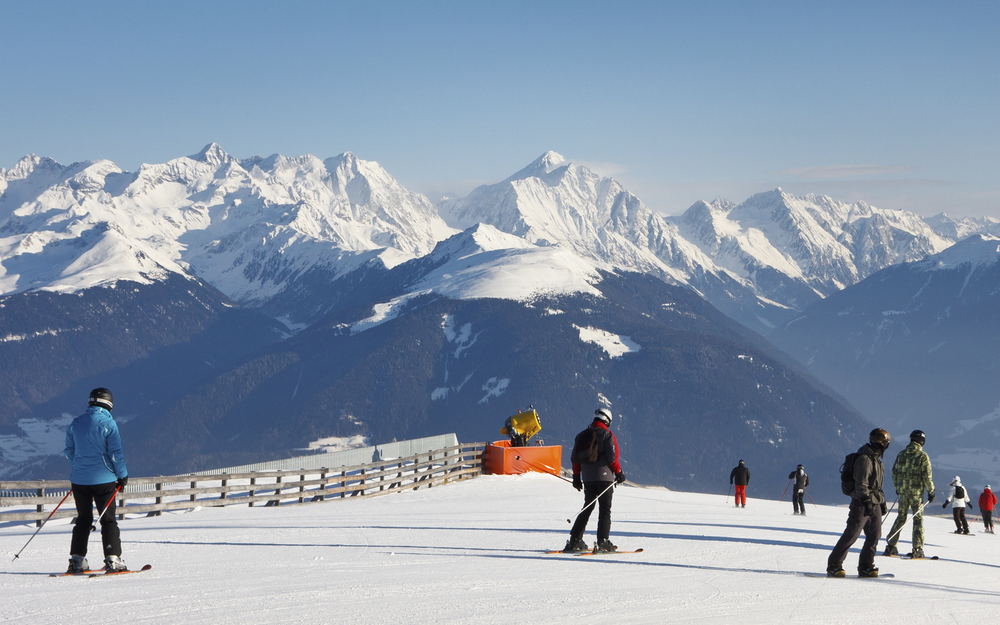 Skipiste am Kronplatz in Südtirol mit Skifahrern, Sonnenschein, blauem Himmel und Blick auf verschneite Berge.