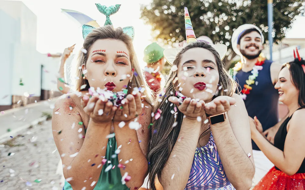 Weiberfastnacht in Köln: verkleidete Frauen (Meerjungfrau, Einhorn) pusten Konfetti, Menschen feiern im Hintergrund.