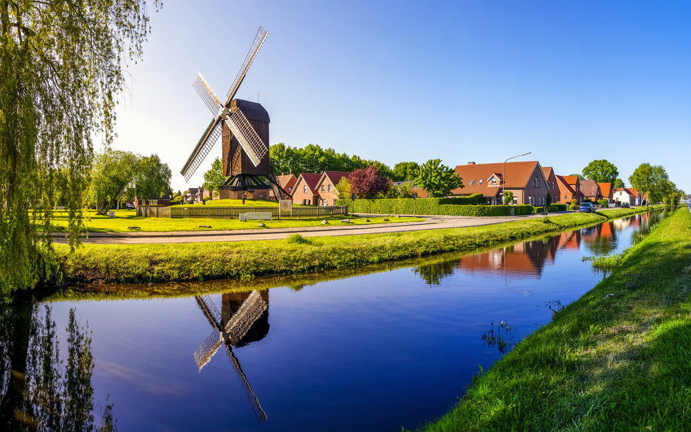 Windmühle neben einem Kanal, spiegelnd im Wasser, vor Häusern und blauem Himmel.