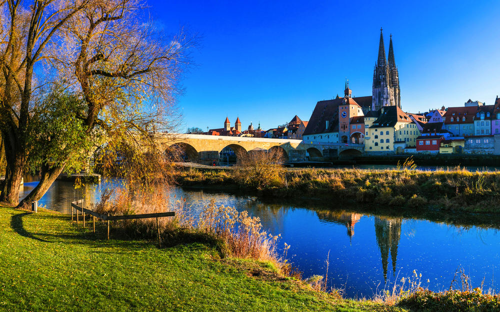 Farbige Stadtansicht mit Fluss und Steinbrücke bei blauem Himmel.