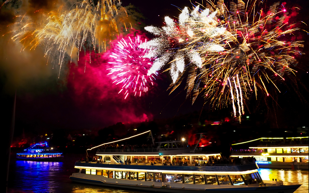 Ein beleuchtetes Boot fährt nachts unter einem Feuerwerk auf einem Fluss.
