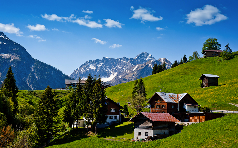 Alpenlandschaft mit Hügeln, Häusern und Bergen im Hintergrund.