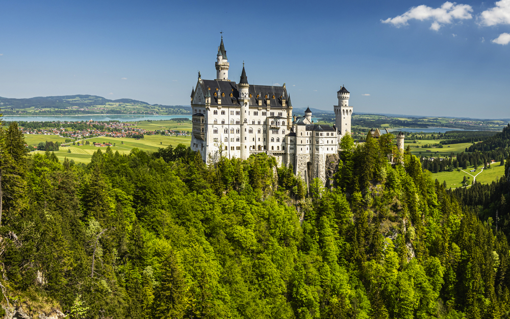 Ein Schloss auf einem bewaldeten Hügel umgeben von üppiger grüner Landschaft unter blauem Himmel.