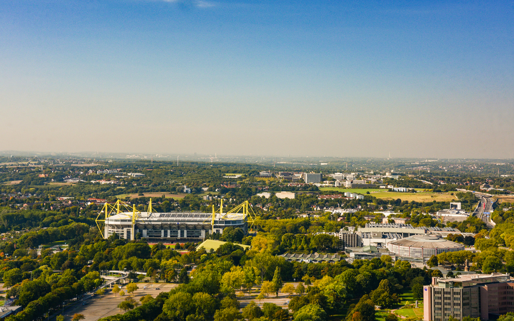 Luftaufnahme eines Stadions inmitten grüner Landschaft und bebauter Umgebung.