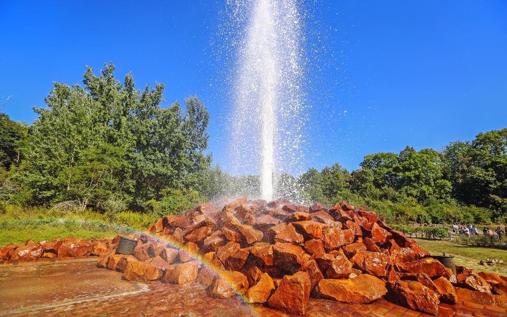 Geysir mit Wasserfontäne über Steinhügel und Regenbogen im Vordergrund.