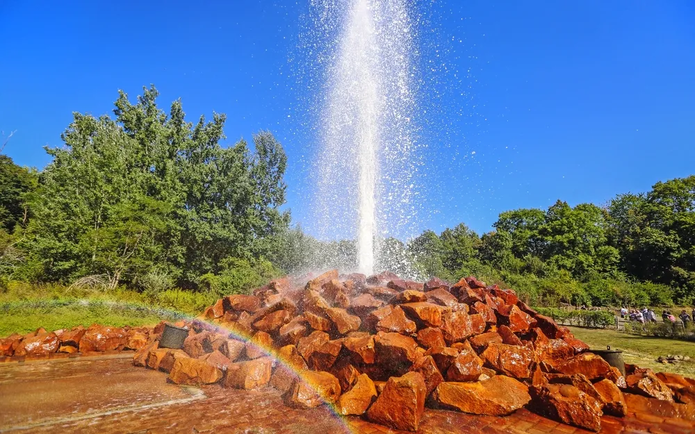Geysir mit Wasserfontäne über Steinhügel und Regenbogen im Vordergrund.