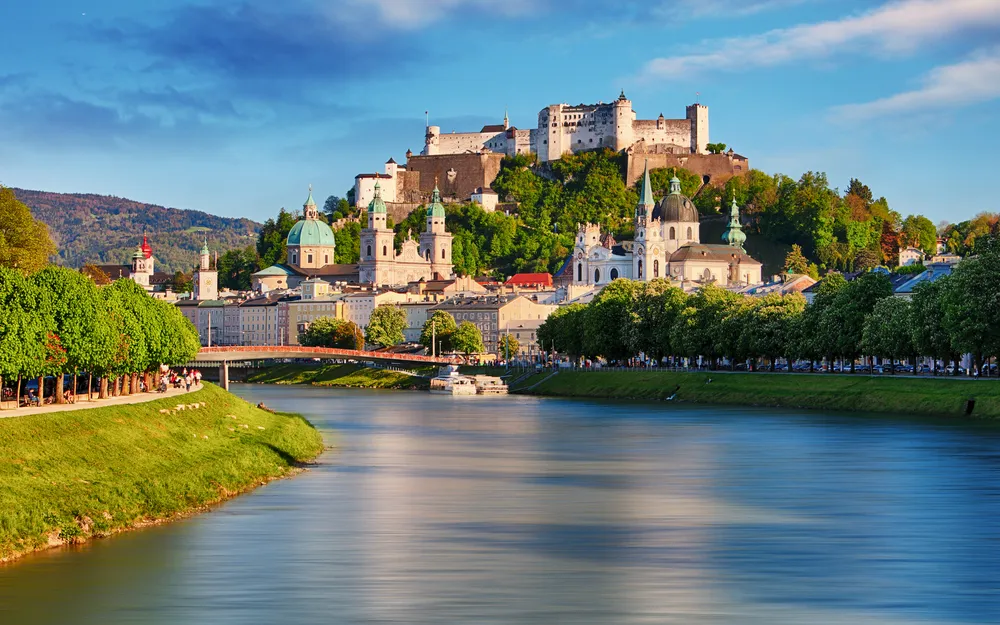 Festung Hohensalzburg über der Stadt Salzburg mit Fluss im Vordergrund bei Sonnenuntergang.