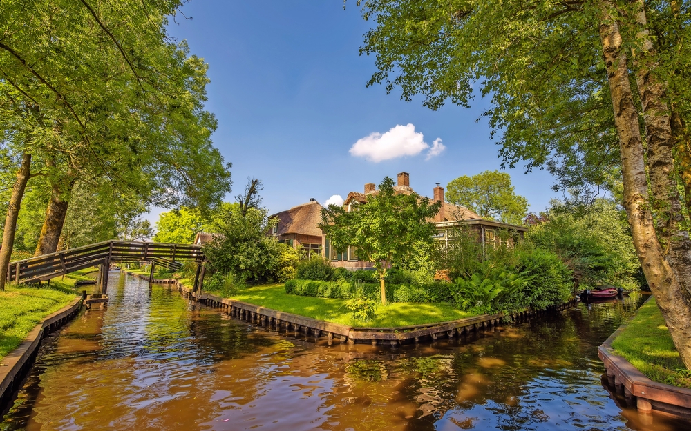 Ein Kanal mit Brücke und umgeben von Bäumen, im Hintergrund ein Haus.