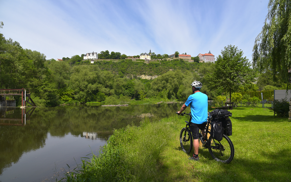 Mann mit Fahrrad am Flussufer, Hügel mit Gebäuden im Hintergrund.