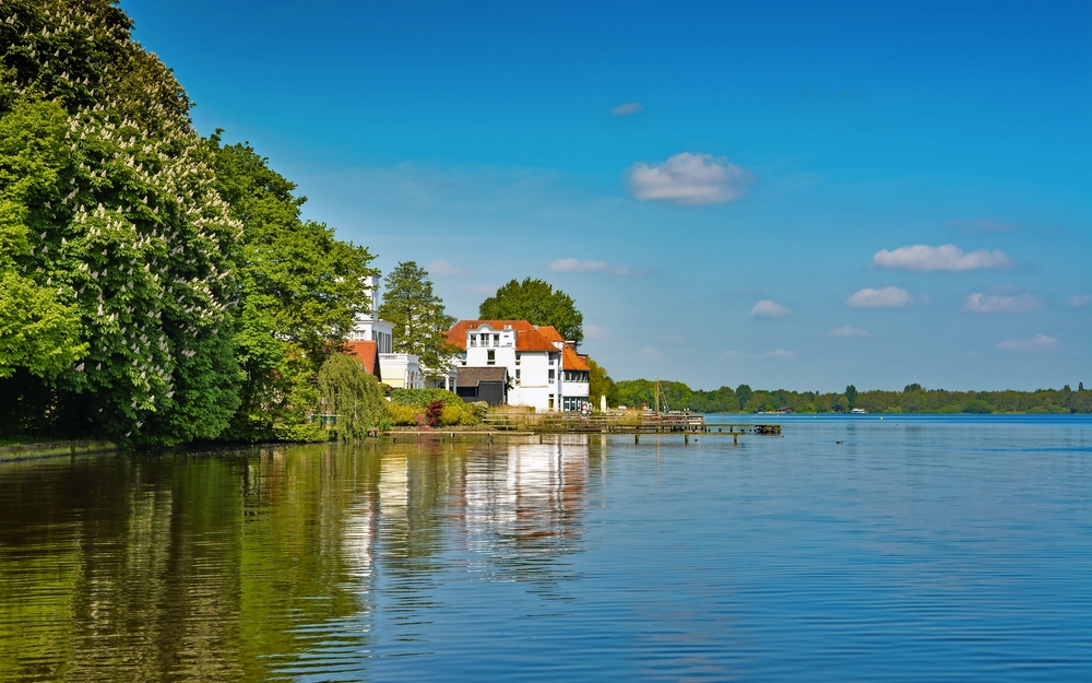 Seeufer mit Häusern und Bäumen unter blauem Himmel.