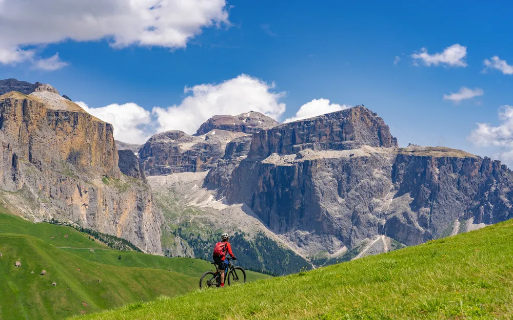 Radfahrer auf grüner Wiese vor Dolomitenpanorama bei klarem Himmel.