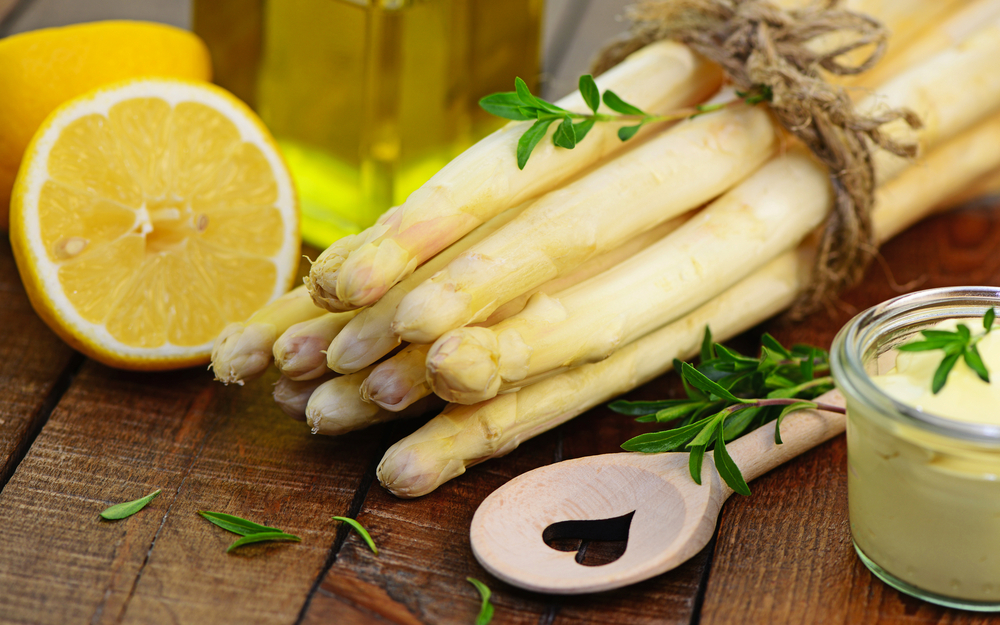 Weiße Spargelstangen mit frischem Zitronenstück und Sauce Hollandaise auf einem Holzbrett.