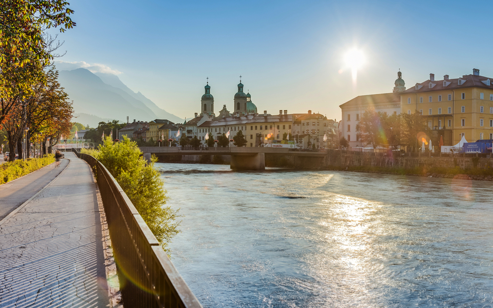 Flussufer mit Stadtansicht bei Sonnenschein und Bergen im Hintergrund.