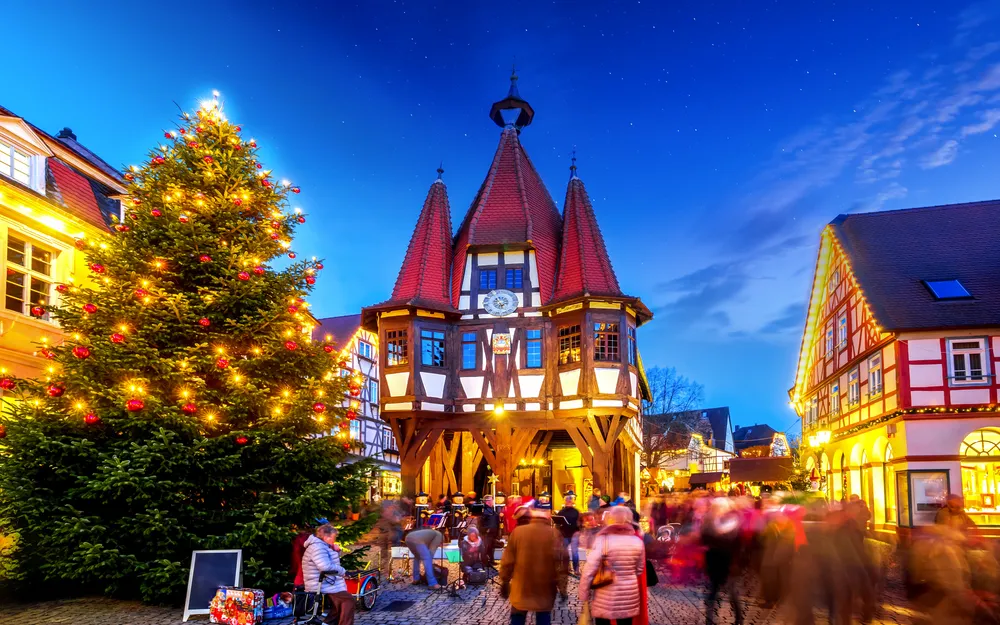 Fachwerkgebäude und Weihnachtsbaum bei Dämmerung in einem geschäftigen Platz.