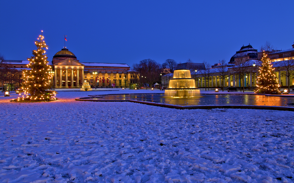 Verschneiter Platz bei Dämmerung mit beleuchteten Gebäuden und Weihnachtsbäumen.