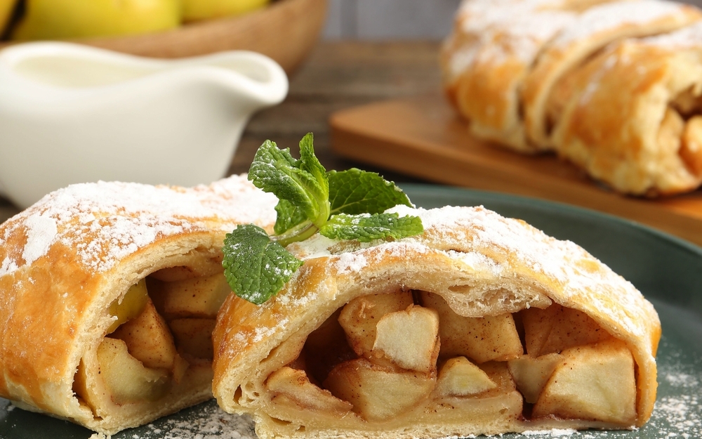 Stücke von leckerem Apfelstrudel mit Puderzucker, Minze und Gabel auf einem Holztisch, Nahaufnahme